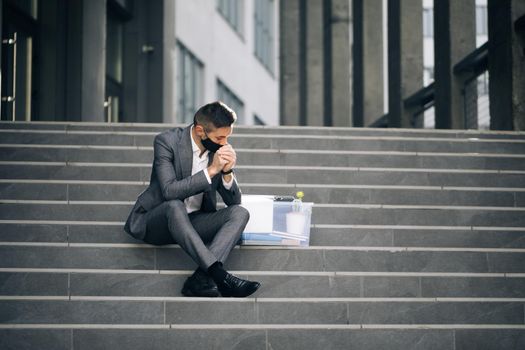 Fired male office worker in medical mask sitting on stairs in depression with box of stuff. Unemployed businessman lost his business. Anxious concept. Workless man in despair.