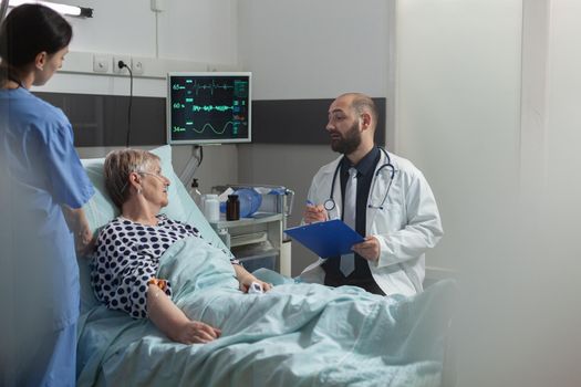 Doctor talking with elderly patient sitting next to bed in hospital room, giving expertise for treatment. Senior woman breathing with help from oxygen mask laying in bed.