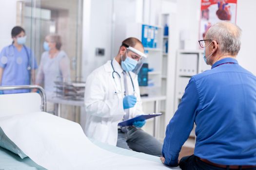 Doctor taking notes on clipboard wearing protective mask against coronavirus outbreak during consultation of senior woman in hospital examination room. Practitioner physician appointment.