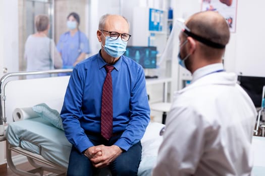Elderly man listening doctor during medical appointment in hospital examination room wearing face mask against coronavirus pandemic. Healthcare medical ,COVID-19 global crisis,
