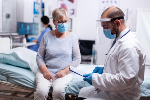 Doctor wearing face mask agasint covid-19 for safety precaution and reading test results to elderly woman in hospital. Healthcare medical physician consultation during COVID-19 global crisis.