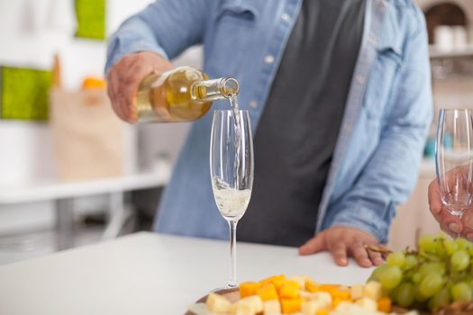 Close up of man pouring sparkling wine in glass on a table top in the kitchen.