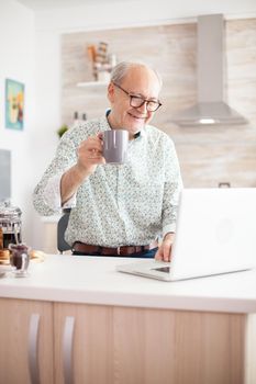 Cheerful senior man during video conference in kitchen on the laptop while enjoying breakfast and a cup of coffee. Elderly person using internet online chat technology video webcam making a video call connection camera communication conference call