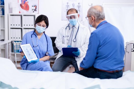 Doctor telling senior man he is suffering of osteoporosis during medical check in hospital room wearing protective mask against covid-19. Medical examination for infections, disease and diagnosis.