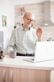 Old man saying hello during video call with family using notebook in kitchen while holding a cup of coffee.