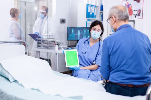 Nurse with face mask for protection against covid 19 holding tablet with green mockup while consulting senior man. Medical examination for infections, disease and diagnosis.