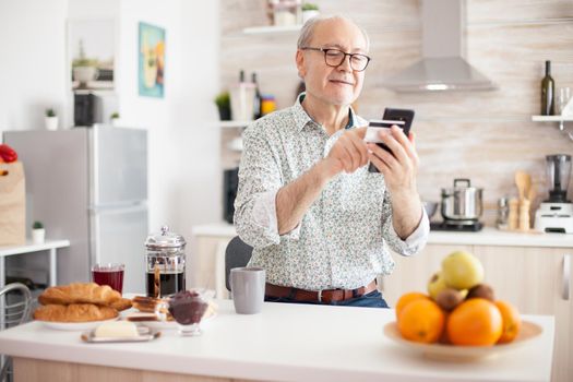 Senior man doing online transaction using phone app for payment during breakfast in kitchen. Retired elderly person using internet payment home bank buying with modern technology