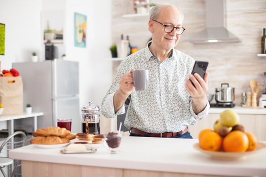 Portrait of elderly man smiling while using modern smartphone in the kitchen. Internet online chat technology video webcam making a video call connection camera communication conference call
