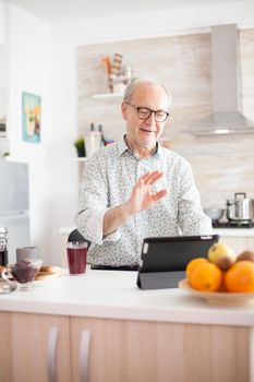 Happy senior man during breakfast in kitchen on a video conference with his family. Elderly person using internet online chat technology video webcam making a video call connection camera communication conference call