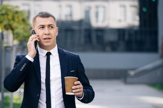 European businessman talking on mobile phone walking near office building background. Man is in dialogue, smiling, holding coffee cup on summer day.