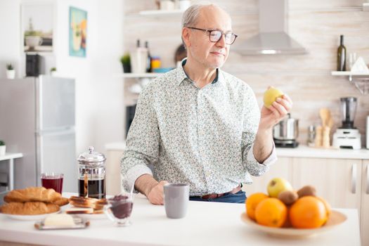 Mature man holding green apple before taking a bite during breakfast in kitchen. Elderly retired person eating apple during breakfast in modern cozy kitchen. Healthy green happy fresh lifestyle