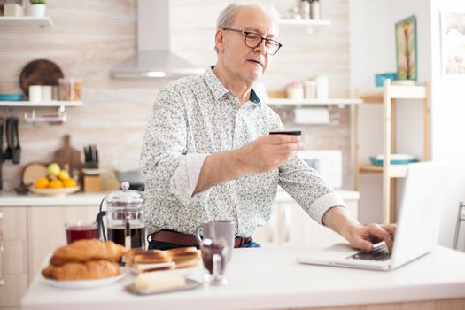 Retired man entering pin number while shopping online on the internet. Pensioner paying online using credit card and application from laptop during breakfast in kitchen. Retired elderly person using internet payment home bank buying with modern technology