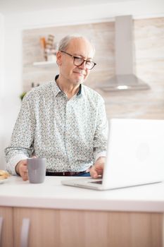 Grayhaired senior retired man browsing on internet. Daily life of senior man in kitchen during breakfast using laptop holding a cup of coffee. Elderly retired person working from home, telecommuting using remote internet job online communication