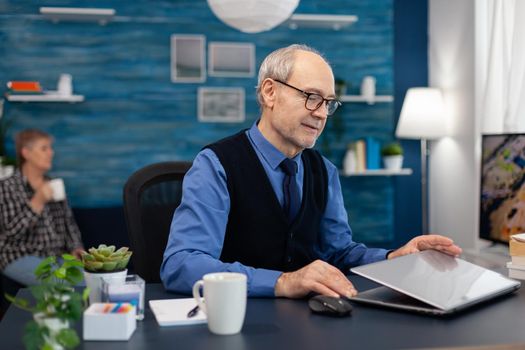 Businessman with gray hair opening laptop to start working. Elderly man entrepreneur in home workplace using portable computer sitting at desk while wife is holding tv remote.