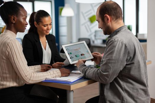 Enthusiastic diverse businesspeople reading annual financial report sitting at table in modern startup business office holding tablet and smiling. Team of multiethnic businesspeople working in company