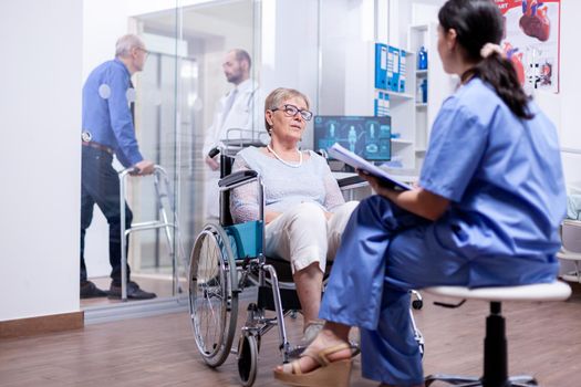 Paralyzed senior woman sitting in wheelchair while having a conversation with nurse dressed in uniform. Help for disabled people. Elderly assistance for old man with walking frame.