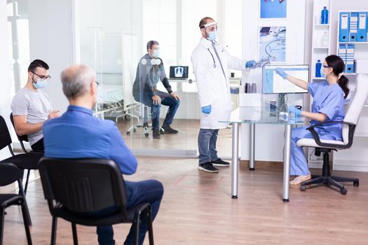 Doctor taking patient x-ray from nurse wearing face mask and visor against cornavirus global pandemic and group of people are waiting for examination. Young man waiting for medic consultation. Health care system.