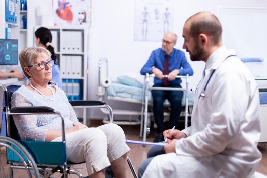 Doctor consultation in recovery clinic of handicapped senior woman in wheelchair. Man with disabilities ,walking frame sitting in hospital bed. Health care system, patients.