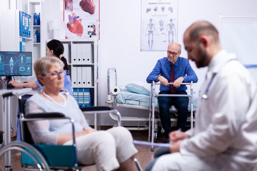 Invalid senior man with walking frame in busy modern private recovery clinic. Man with disabilities ,walking frame sitting in hospital bed. Health care system, patients.