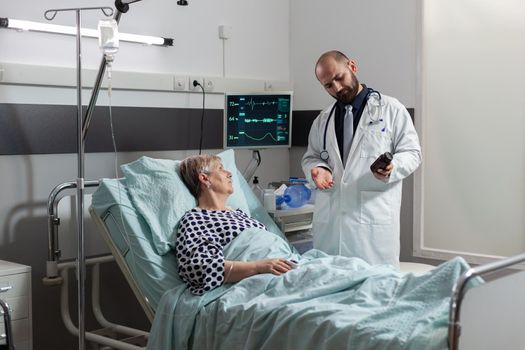 Senior woman patient breathing with help from oxygen tube laying in hospital bed, listening doctor explaying diagnosis treatmant holding pills bottle for recovery.