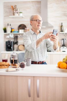 Mature man surfing the social media using phone during breakfast in kitchen. Elderly person using internet online chat technology video webcam making a video call connection camera communication conference call