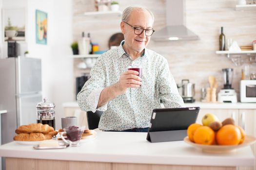 Elderly man using tablet pc in kitchen during breakfast wearing glasses. Elderly person with tablet portable pad PC in retirement age using mobile apps, modern internet online information technology with touchscreen