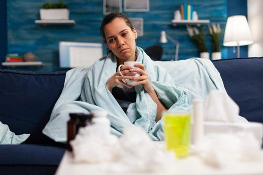 Sick caucasian woman sitting in bed with severe cold and flu drinking hot tea. Adult wrapped in blanket on the sofa with health fever infection symptomps. Person with disease at home, temperature and cough