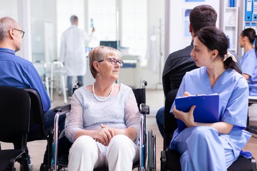 Nurse filing documents while talking with disabled senior woman in hospital waiting area. Patient asking for direction at hospital reception.