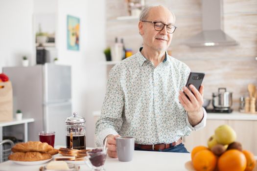 Senior man browsing on internet using smartphone in kitchen while enjoying morning coffee during breakfast. Authentic portrait of retired senior enjoying modern internet online technology