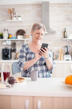 Elderly woman reading message on smartphone during breakfast in kitchen. Authentic elderly person using modern smartphone internet technology. Online communication connected to the world, senior leisure time with gadget