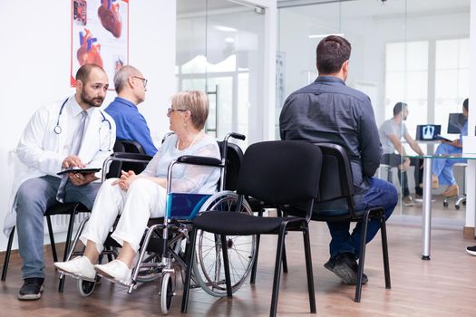 Doctor holding tablet pc with x-ray while explaining diagnosis to disabled elderly woman in wheelchair. Handicapped patient in hospital waiting area. Man in examination room.