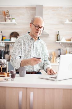 Elderly man holding credit card, using modern payment system. Pensioner paying online using credit card and application from laptop during breakfast in kitchen. Retired elderly person using internet payment home bank buying with modern technolog