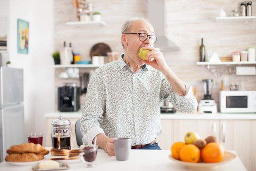 Elderly retired person biting apple during breakfast in mdoern cozy kitchen. Healthy green happy fresh lifestyle with organic food, vitamines, grandpa,