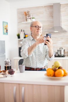 Senior man shopping online using smartphone and holding credit card during breakfast in kitchen. Retired elderly person using internet payment home bank buying with modern technology