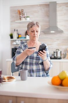 Senior woman paying online using phone app and holding credit card in kitchen during breakfast. Retired elderly person using internet payment home bank buying with modern technology