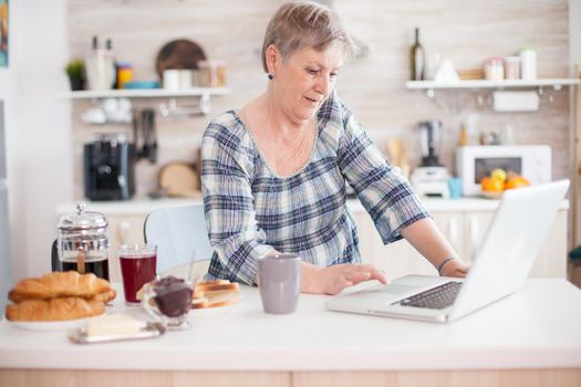 Senior woman drinking coffee and working on laptop in kitchen during breakfast. Elderly retired person working from home, telecommuting using remote internet job online communication on modern technology notebook
