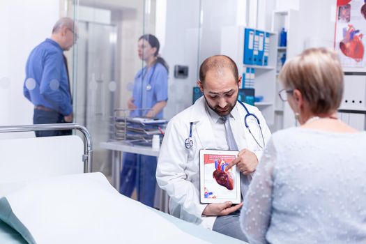 Doctor explaining coronary artery disease to senior woman patient pointint at tablet pc in hospital examination room. Medical services medicine healthcare well-being support, physician working