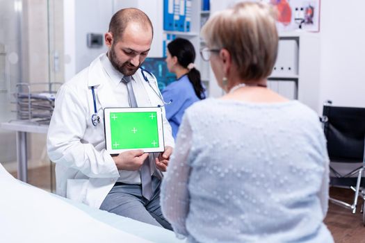 Medical practitioner with green screen tablet in private clinic during consultation of mature patient. Ready chroma mockup for your app, text, video or other digital asset.