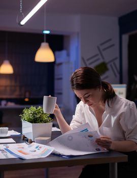 Overworked businesswoman working overtime in business company office meeting room in evening analyzing marketing charts while drinking coffee. Entrepreneur hardworking at financial profit reports