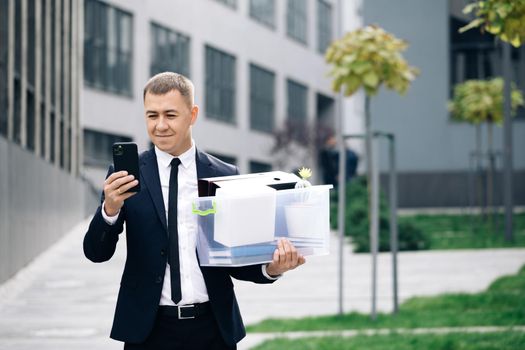 Young businessman with box of personal stuff uses phone texting scrolling tapping. Happy businessman stand smiling use phone near business center. Portrait suit career male office handsome technology.