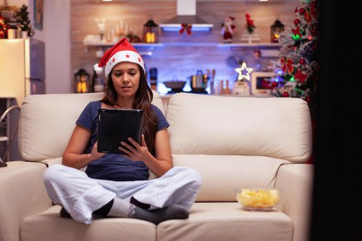 Adult relaxing in xmas decorated kitchen sitting in lotus position on couch browsing social media using tablet computer. Woman with santa hat searching x-mas holiday celebrating winter season