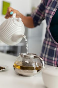 Wife holding teapot with hot water and preparing tea with aromatic herbs in kitchen. Woman, lifestylem, beverage, preparation, herbal, teapot, morning, aromatic.