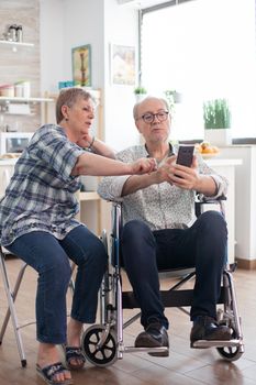 Disabled senior man in wheelchair and his wife are browsing on internet using smartphone. Paralysed man using modern technology. Wife talking with husband.