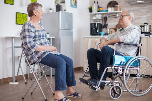 Old woman and her disabled husband in wheelchair chatting in kitchen.Elderly person having a conversation with husband in kitchen. Living with disabled person with walking disabilities
