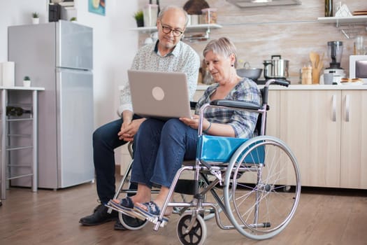 Disabled senior woman in wheelchair and her husband having a video conference on tablet pc in kitchen. Paralyzed old woman and her husband having a online conference.