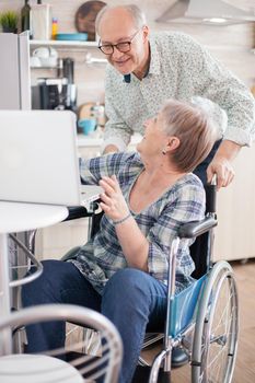 Senior couple laughing during a video call with grandchildrens using tablet computer in kitchen. Paralyzed handicapped old elderly woman using modern communication techonolgy.
