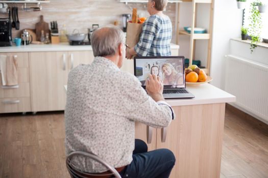 Happy senior man waving to his niece during a video conference with family using laptop in kitchen. Online call with daughter. Elderly person using modern communication online internet web techonolgy.