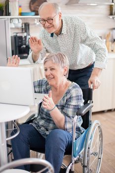 Senior couple laughing and waving during a video call with grandchildrens using tablet computer in kitchen. Paralyzed handicapped old elderly man using modern communication techonolgy.
