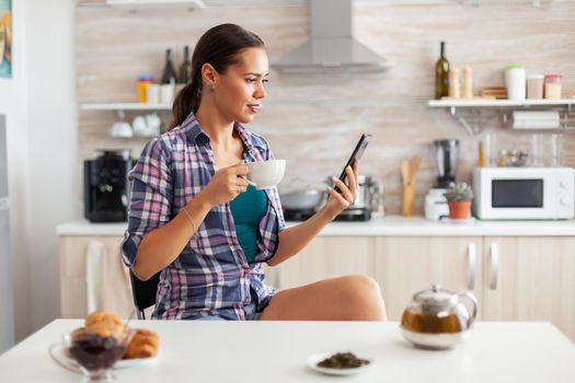 Woman holding phone and drinking hot tea with aromatic herbs in kitchen during breakfast. Holding phone device with touchscreen using internet technology scrolling, searching on intelligent gadget.