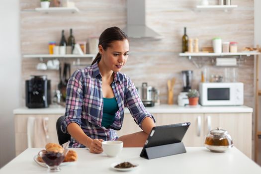 Cheerful woman smiling and reading news on tablet while drinking green tea. Working from home using device with internet technology, typing, on gadget during breakfast.